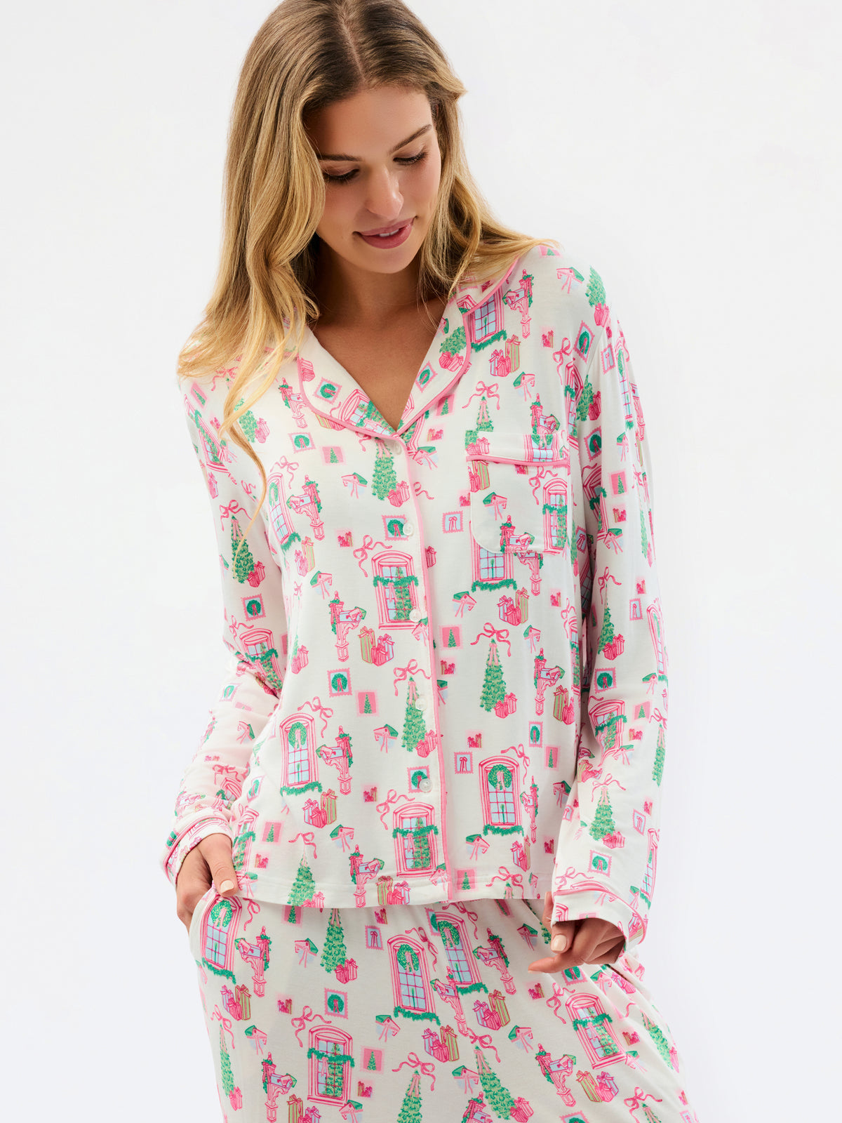 A woman wearing a matching long-sleeve pajama top and pants set with a whimsical pink and green house pattern on a cream background, photographed against a plain white studio backdrop.