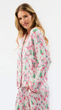 A woman modeling a matching pajama set with a long-sleeve button-front top and coordinating pants, featuring a pink and green gingerbread house and tree pattern on a light cream background.