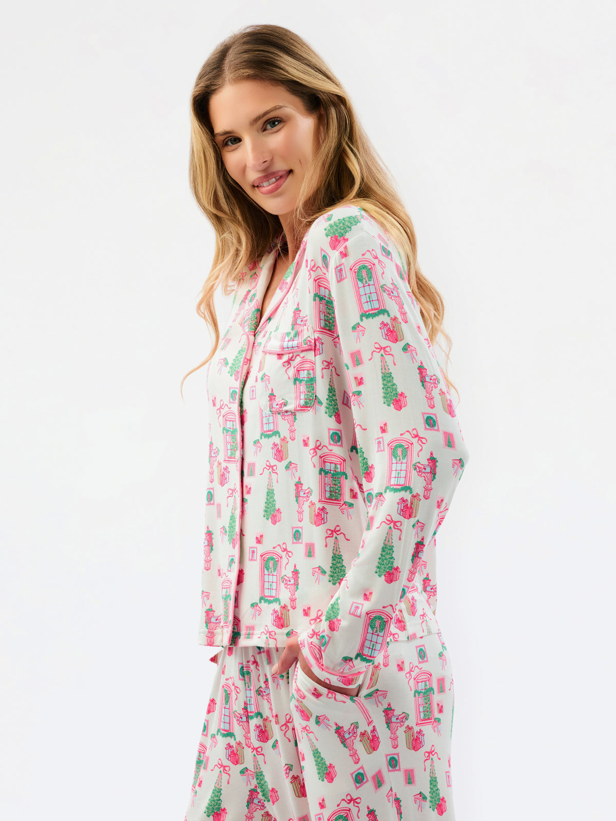 A woman modeling a matching pajama set with a long-sleeve button-front top and coordinating pants, featuring a pink and green gingerbread house and tree pattern on a light cream background.