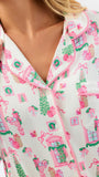 A close-up of a person wearing a pajama top with a festive holiday village print. The pajamas are off-white with pink piping and an all-over pattern of pink houses, green wreaths, Christmas trees, and pink bows.