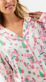 A close-up of a woman wearing a long-sleeve pajama top with a festive holiday village print in pink and green on a white background.