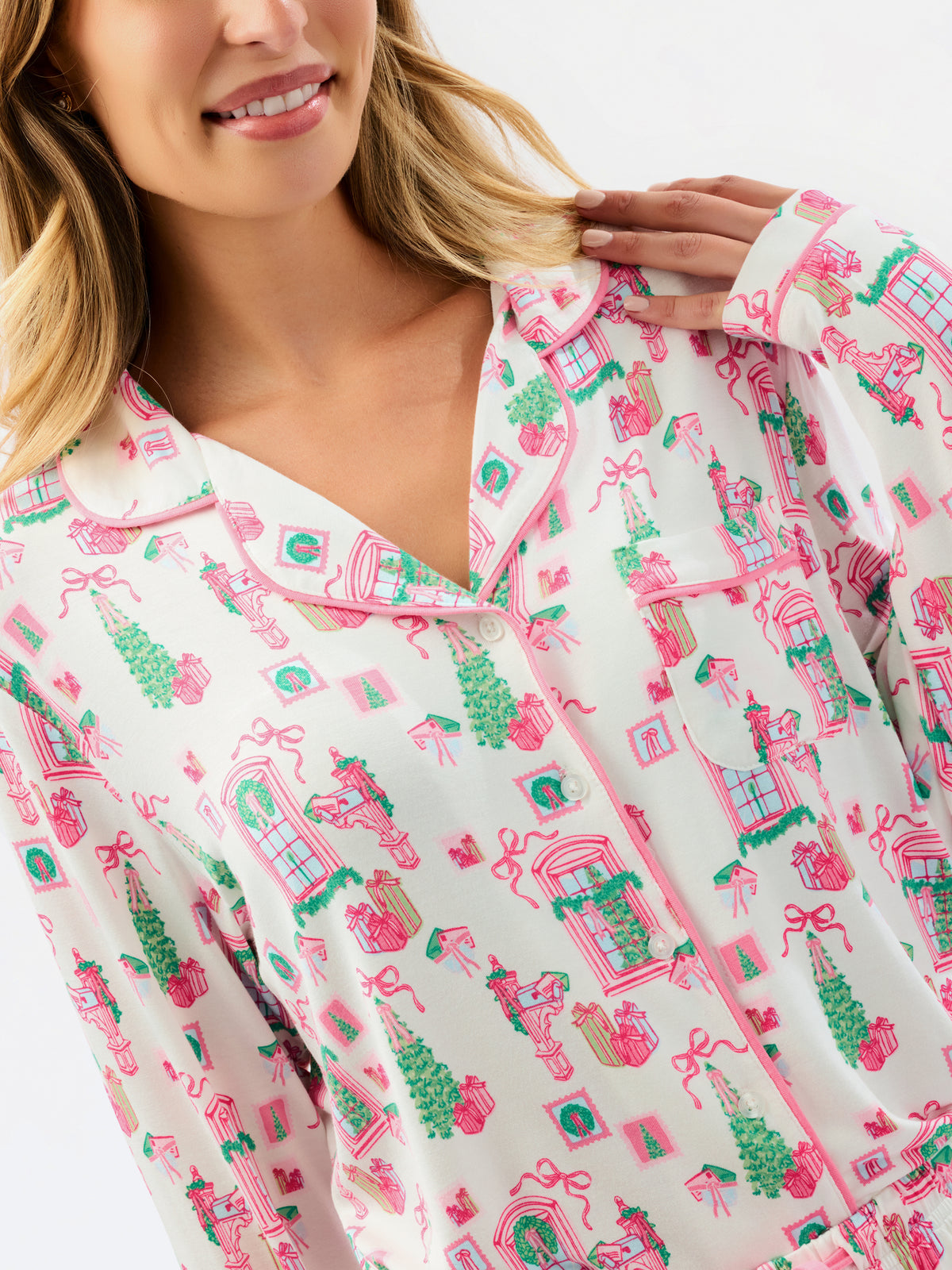 A close-up of a woman wearing a long-sleeve pajama top with a festive holiday village print in pink and green on a white background.