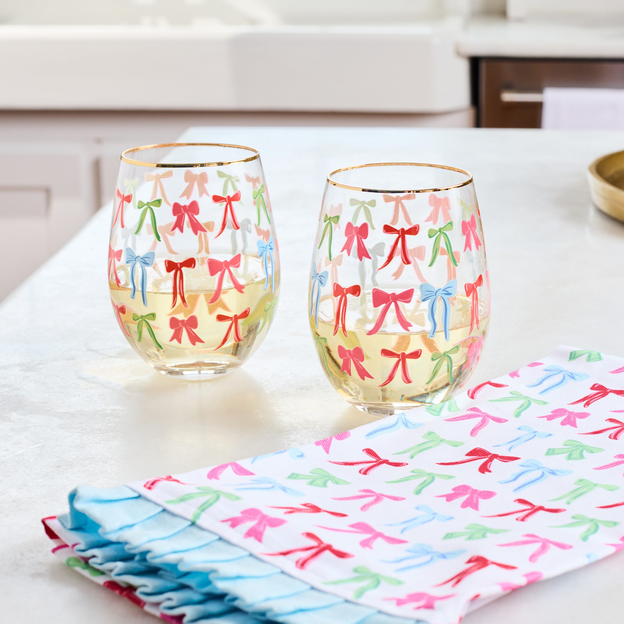 Two clear stemless wine glasses with gold rims decorated with colorful bow patterns, set on a white countertop next to a matching folded cloth napkin with ruffled edges.