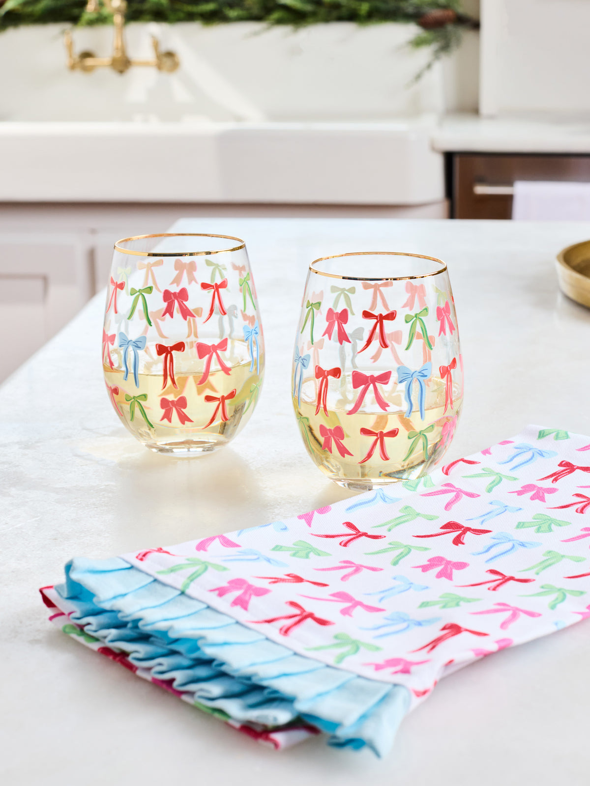 Two clear stemless wine glasses with gold rims decorated with colorful bow patterns, set on a white countertop next to a matching folded cloth napkin with ruffled edges.