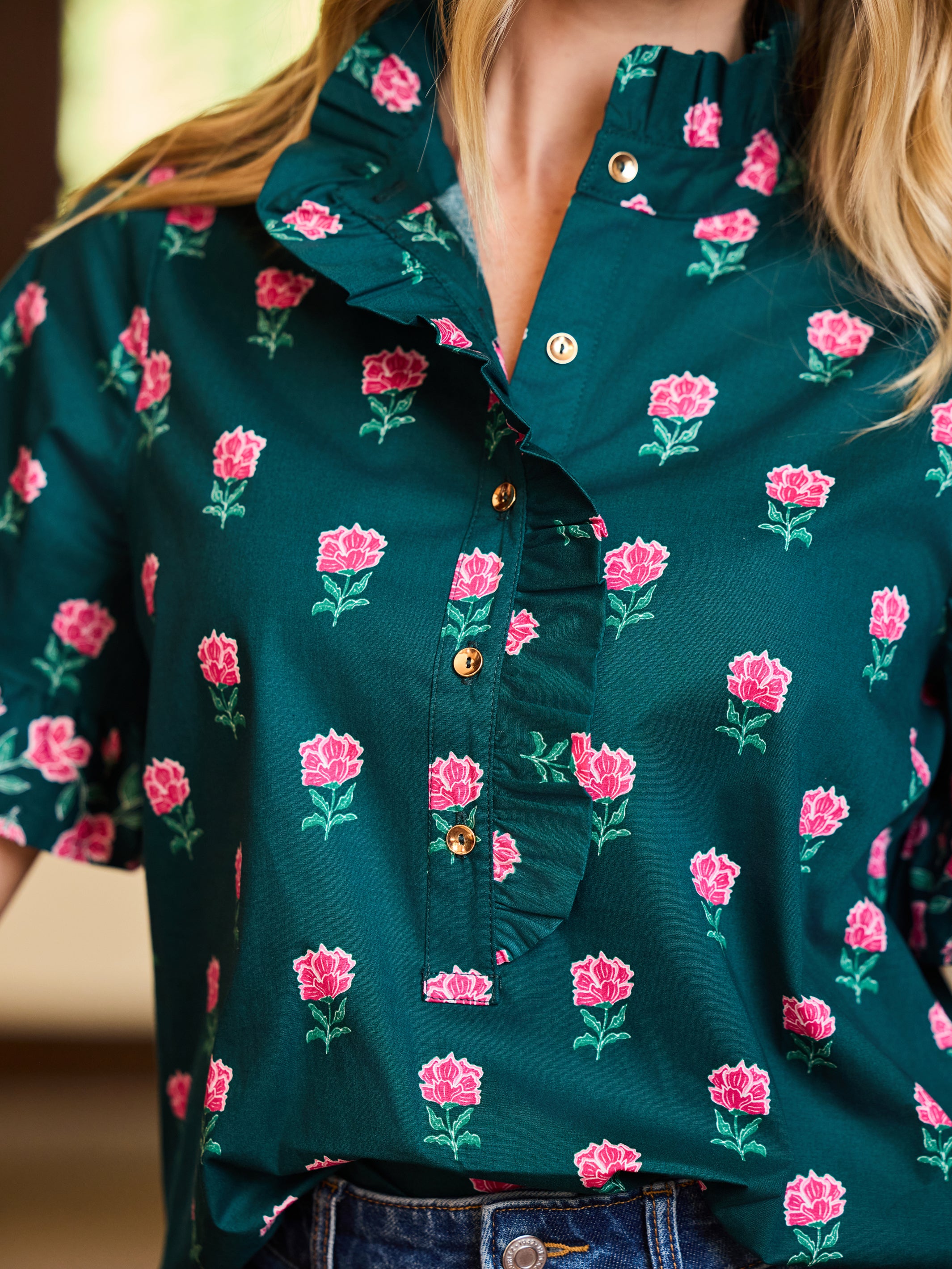 A close-up of a woman wearing a dark teal green short-sleeve blouse with a pink rose floral print and gold buttons, paired with blue jeans.