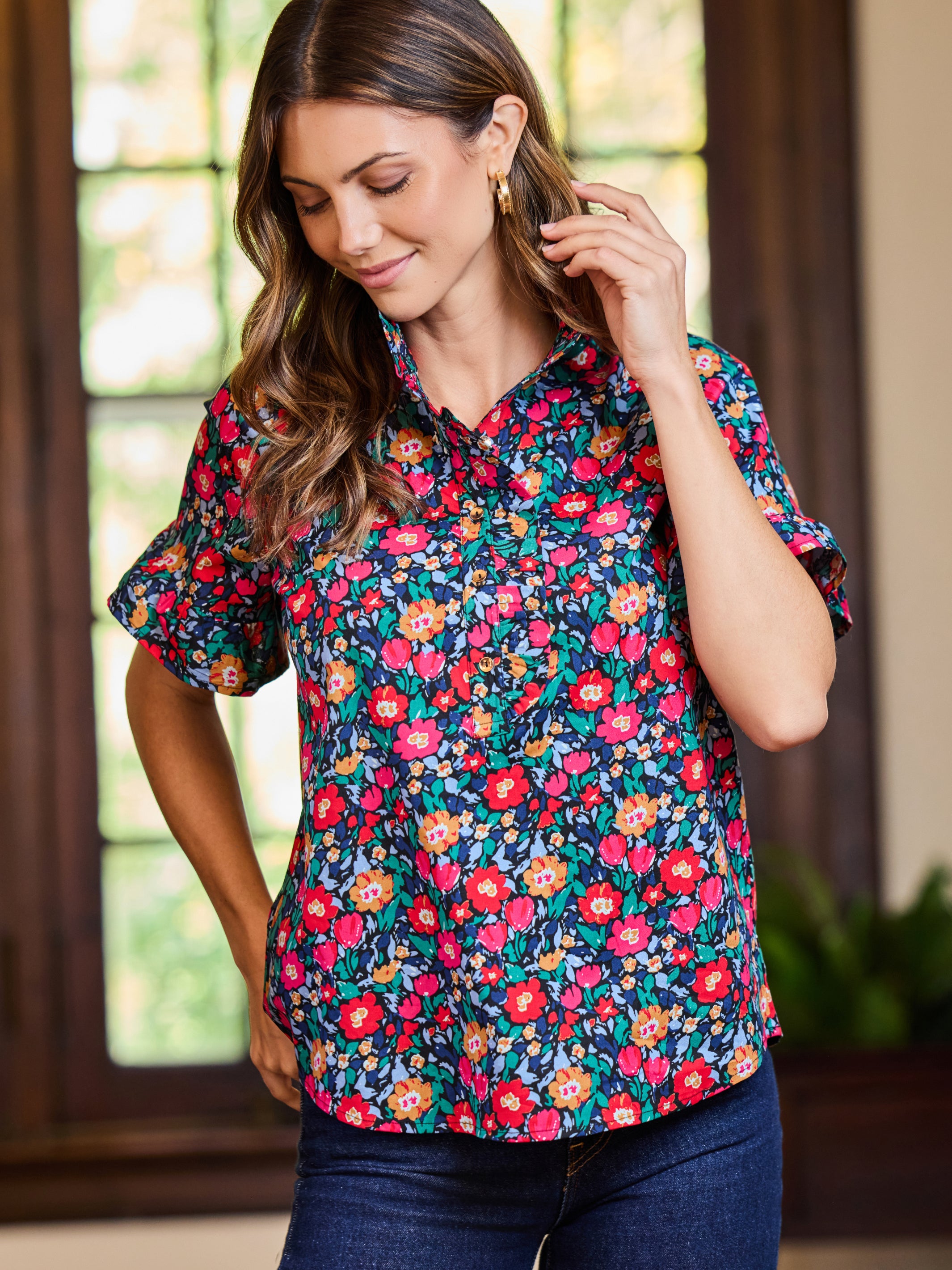 A woman wearing a short-sleeve button-up blouse with a vibrant multicolor floral print, paired with dark blue jeans, standing in front of a softly blurred window.