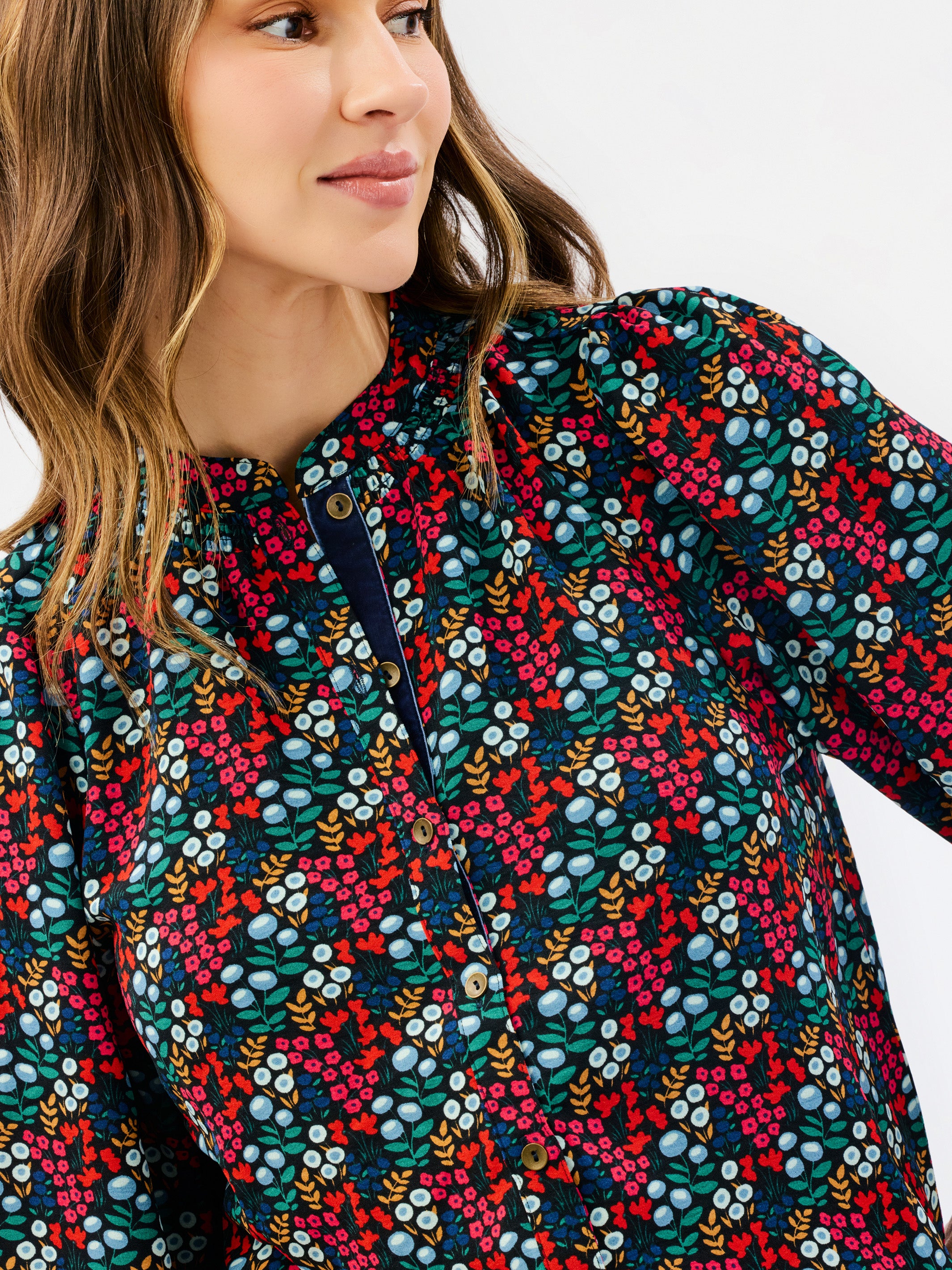 A close-up of a woman wearing a long-sleeve button-up shirt with a dense, colorful floral and berry print on a dark background.
