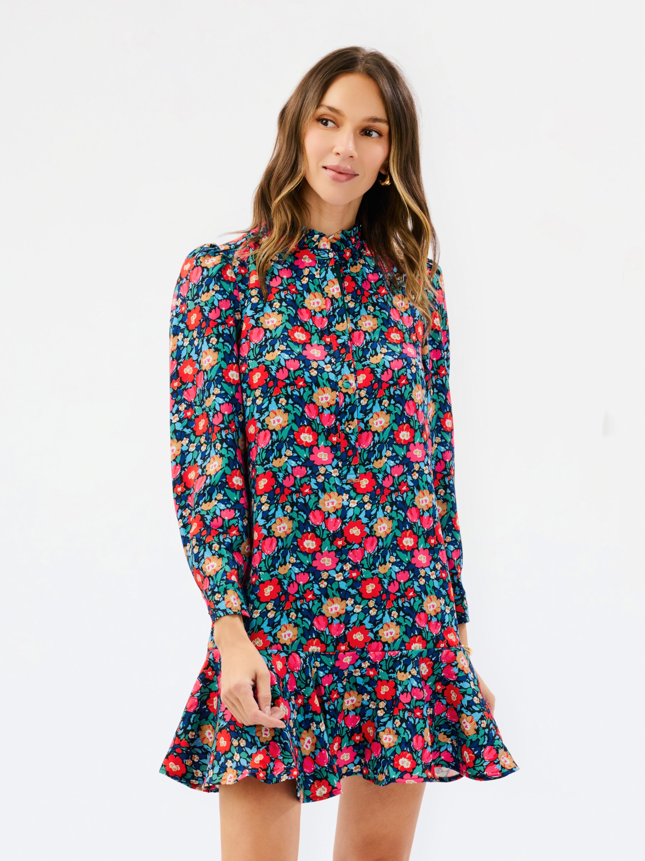 A woman modeling a long-sleeve floral dress with a high gathered neckline and a ruffled hem, set against a plain white studio background.
