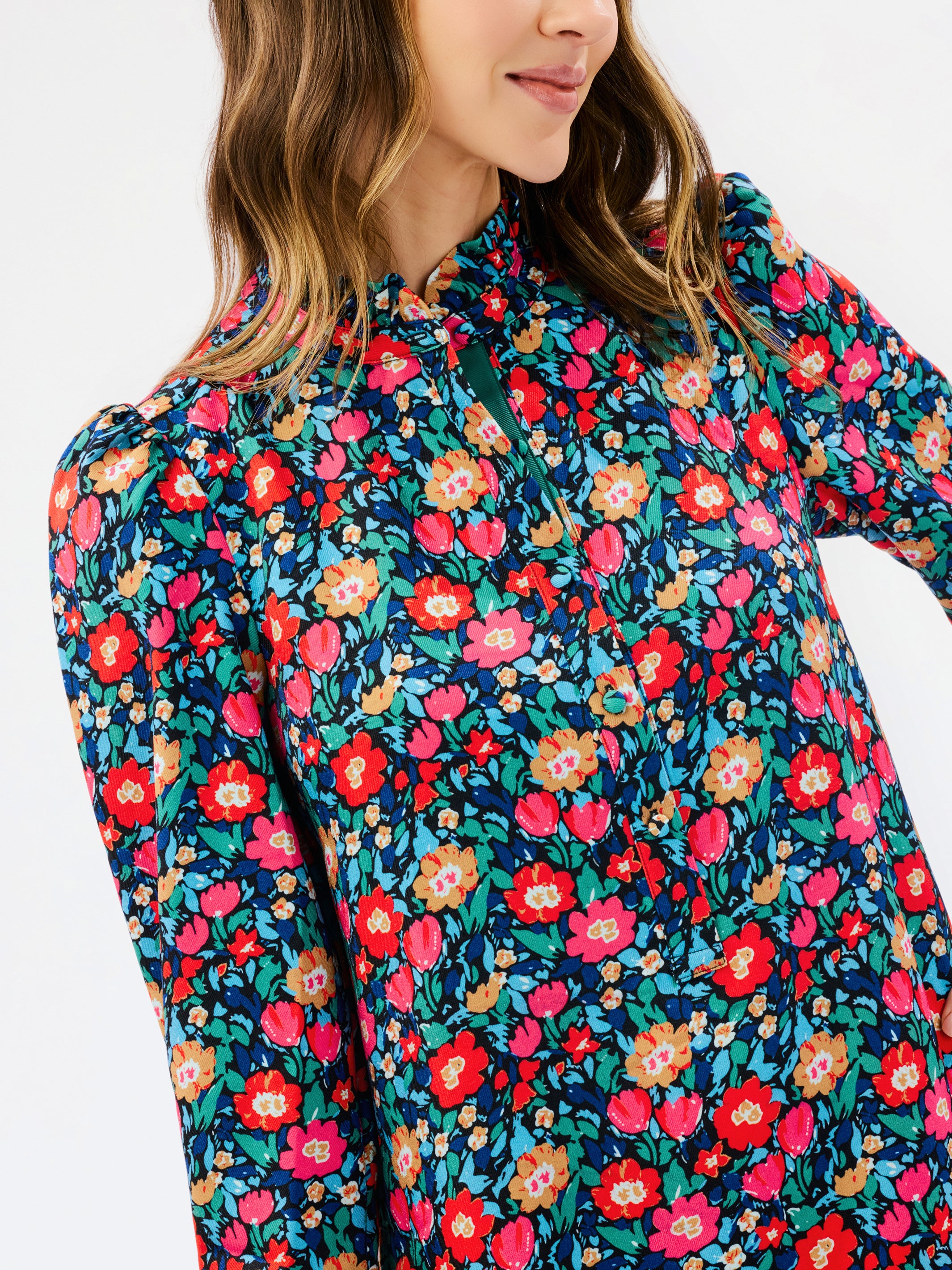 A close-up of a woman wearing a long-sleeve floral print top with a high mock neck. The top features a dense pattern of red, pink, orange, and yellow flowers with green leaves on a dark teal background.