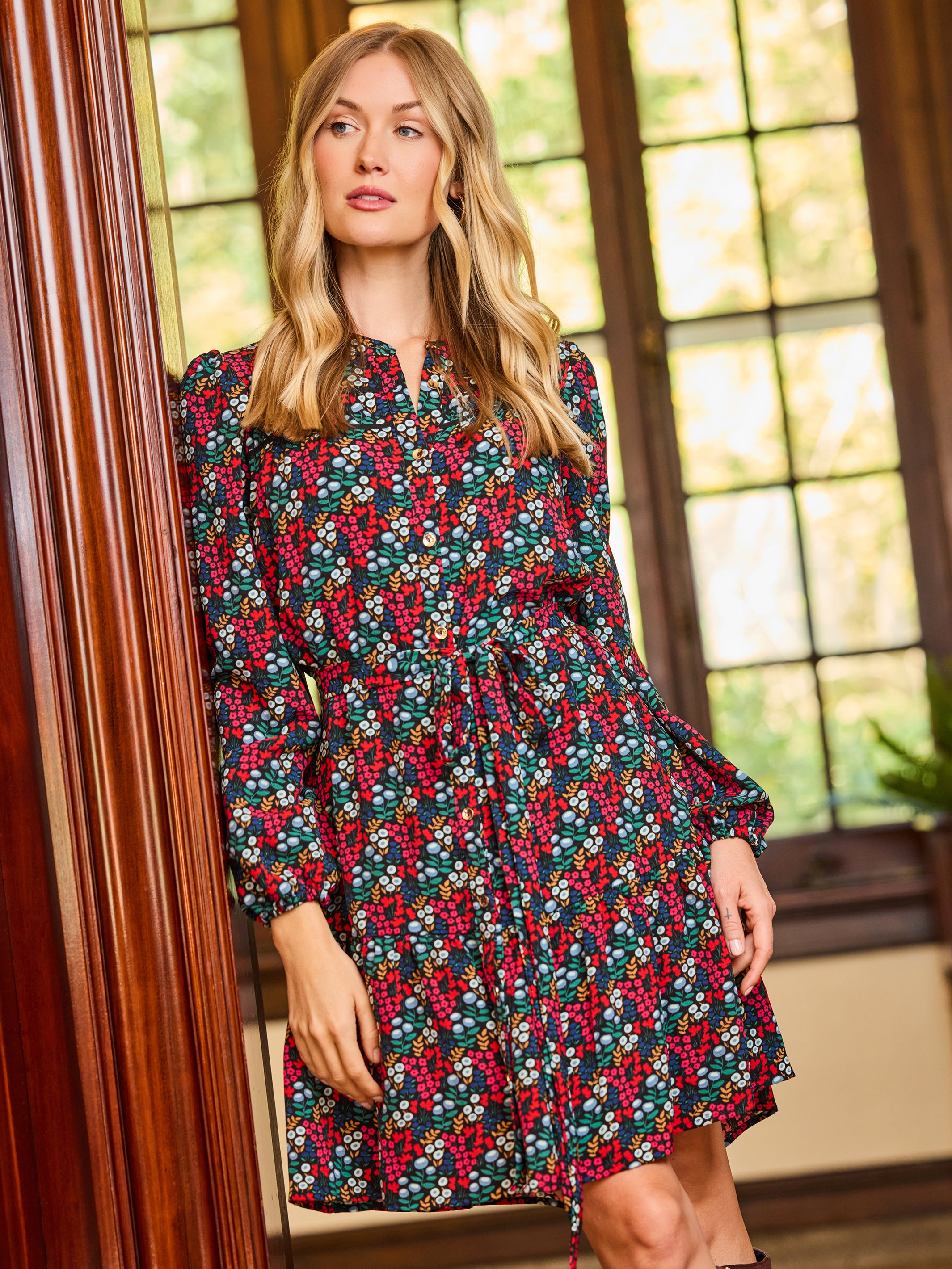 A woman wearing a long-sleeve floral dress with a red, green, and white berry pattern, standing in a room with large wooden-framed windows.