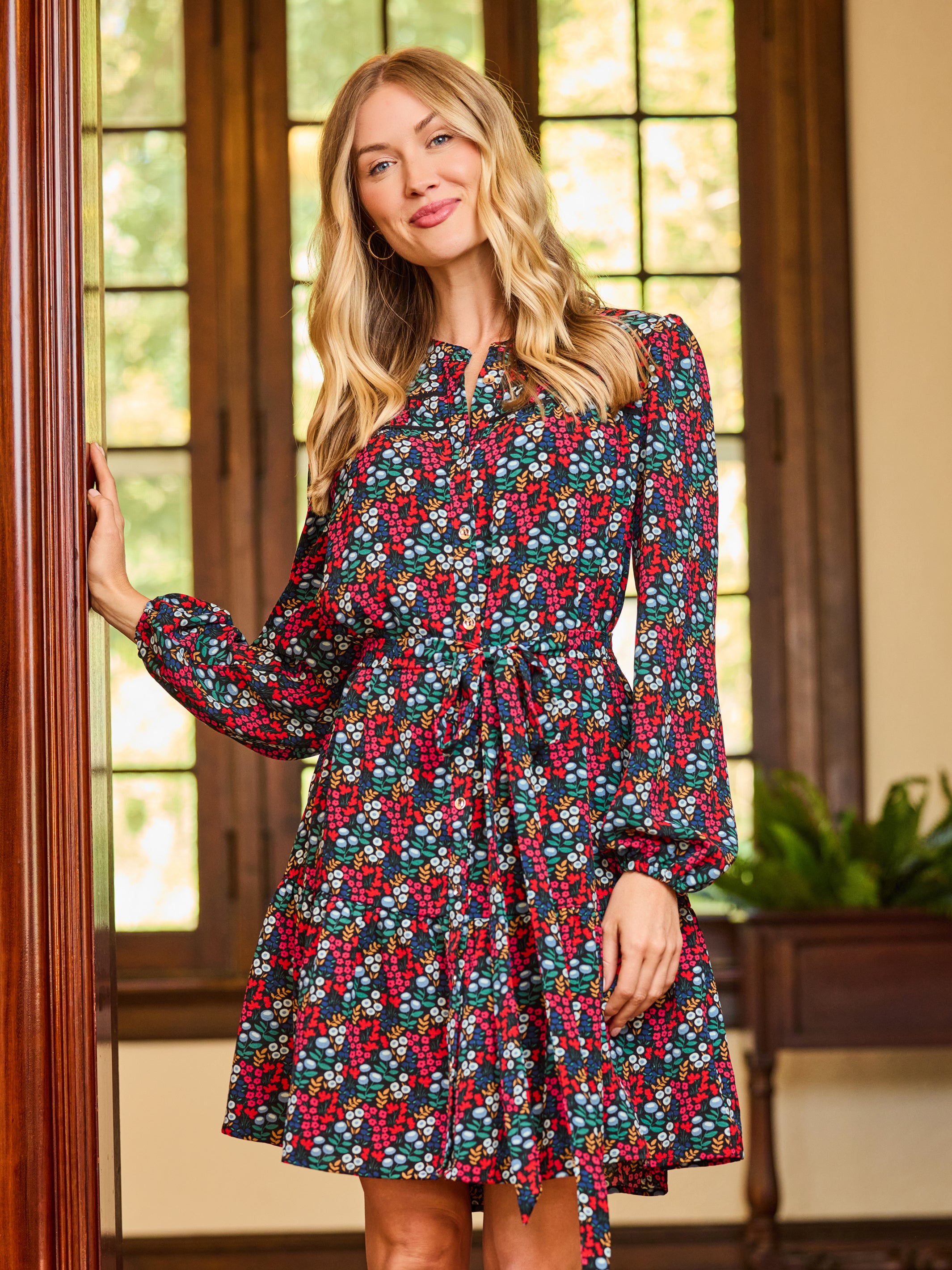 A woman wearing a long-sleeve floral dress with a red, green, and white berry pattern, standing in a warm indoor setting with wooden window frames.