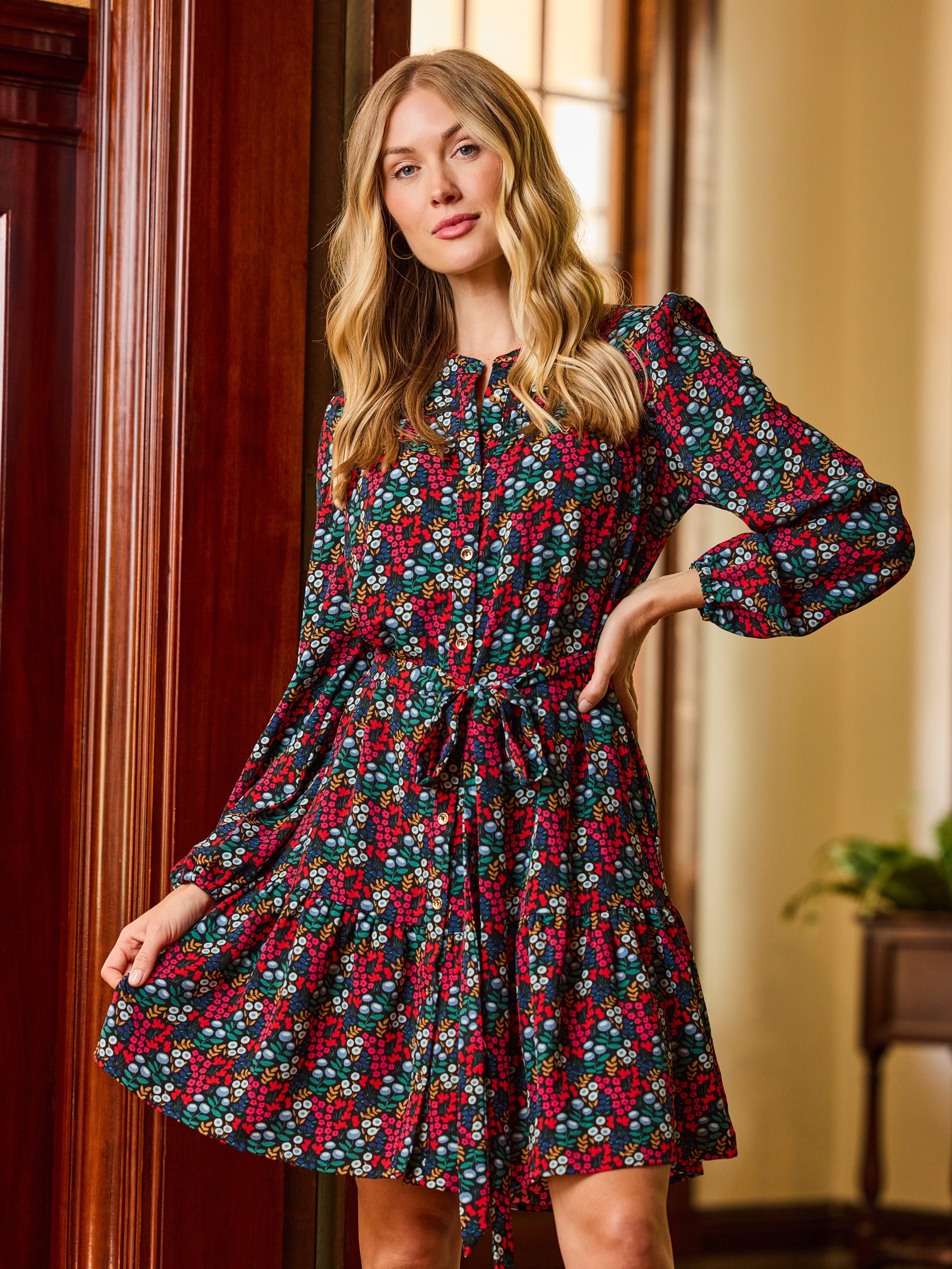A woman modeling a long-sleeve, tiered mini dress with a red and white floral print on a dark green background, standing in a warm-toned indoor setting.
