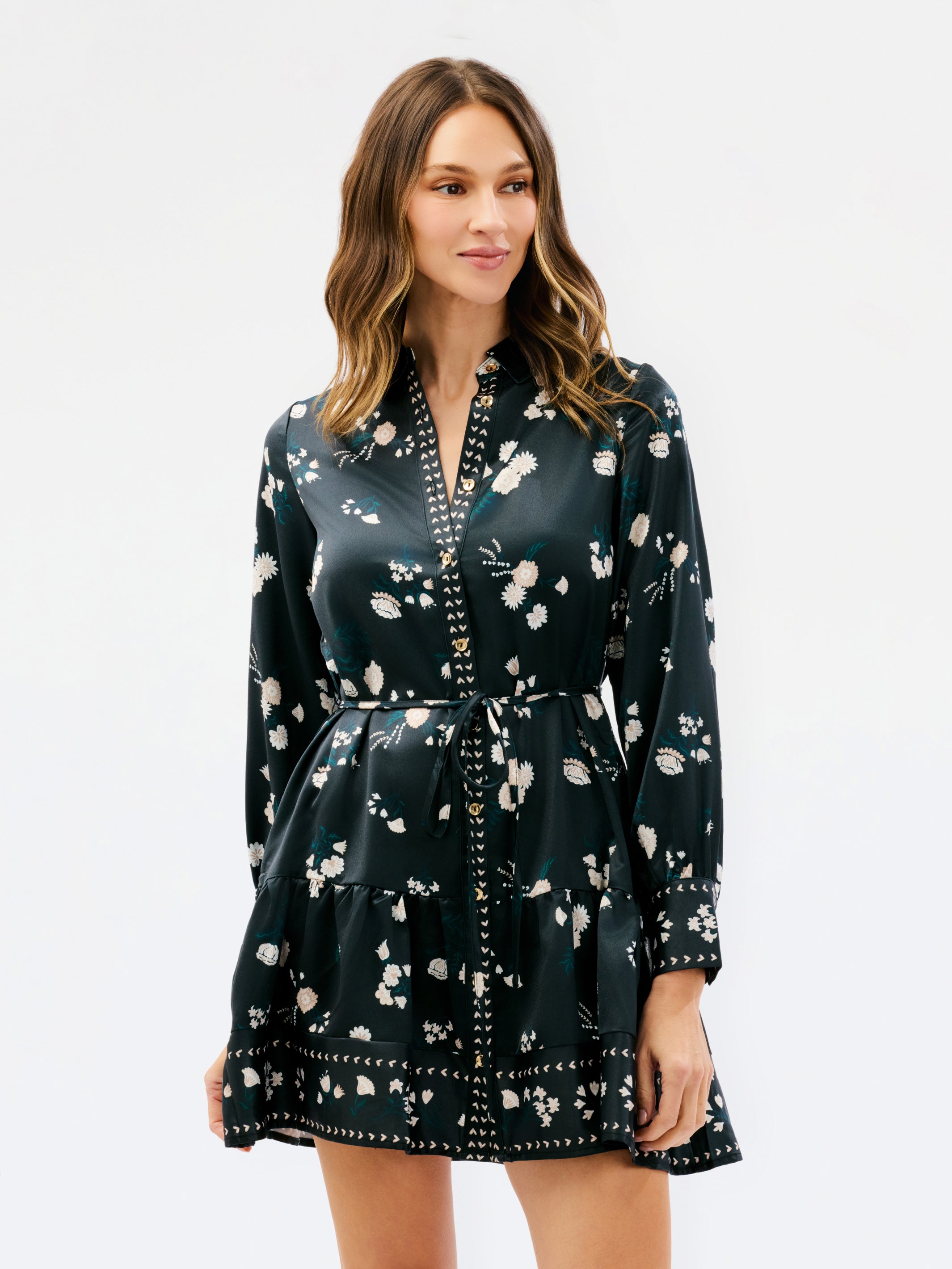 A woman modeling a dark green satin-like mini dress with a white floral print, a V-neckline, and a matching tie belt, set against a plain white studio background.