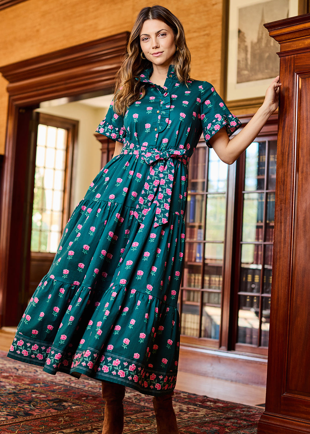 A woman models a dark teal green midi dress with a pink and white floral print, featuring a button-up front, short flutter sleeves, and a matching fabric tie belt at the waist. She is standing in a warm, wood-paneled room with bookshelves in the background.