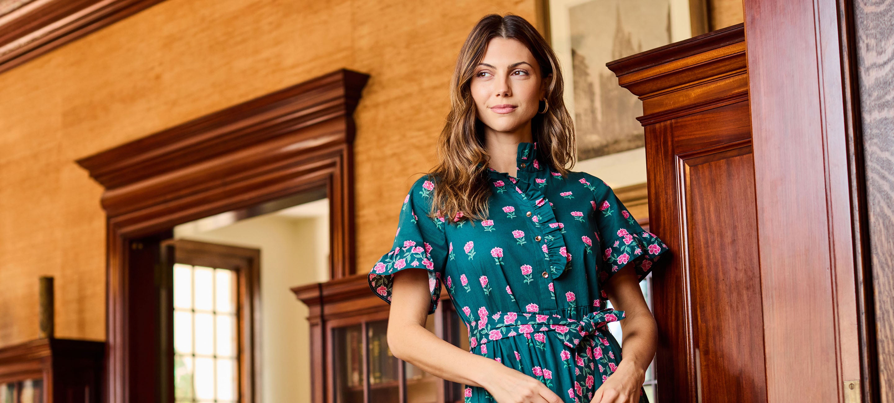 A woman wearing a teal green short-sleeve dress with a pink and white floral pattern, standing in a room with warm wood-paneled walls and wooden doors.