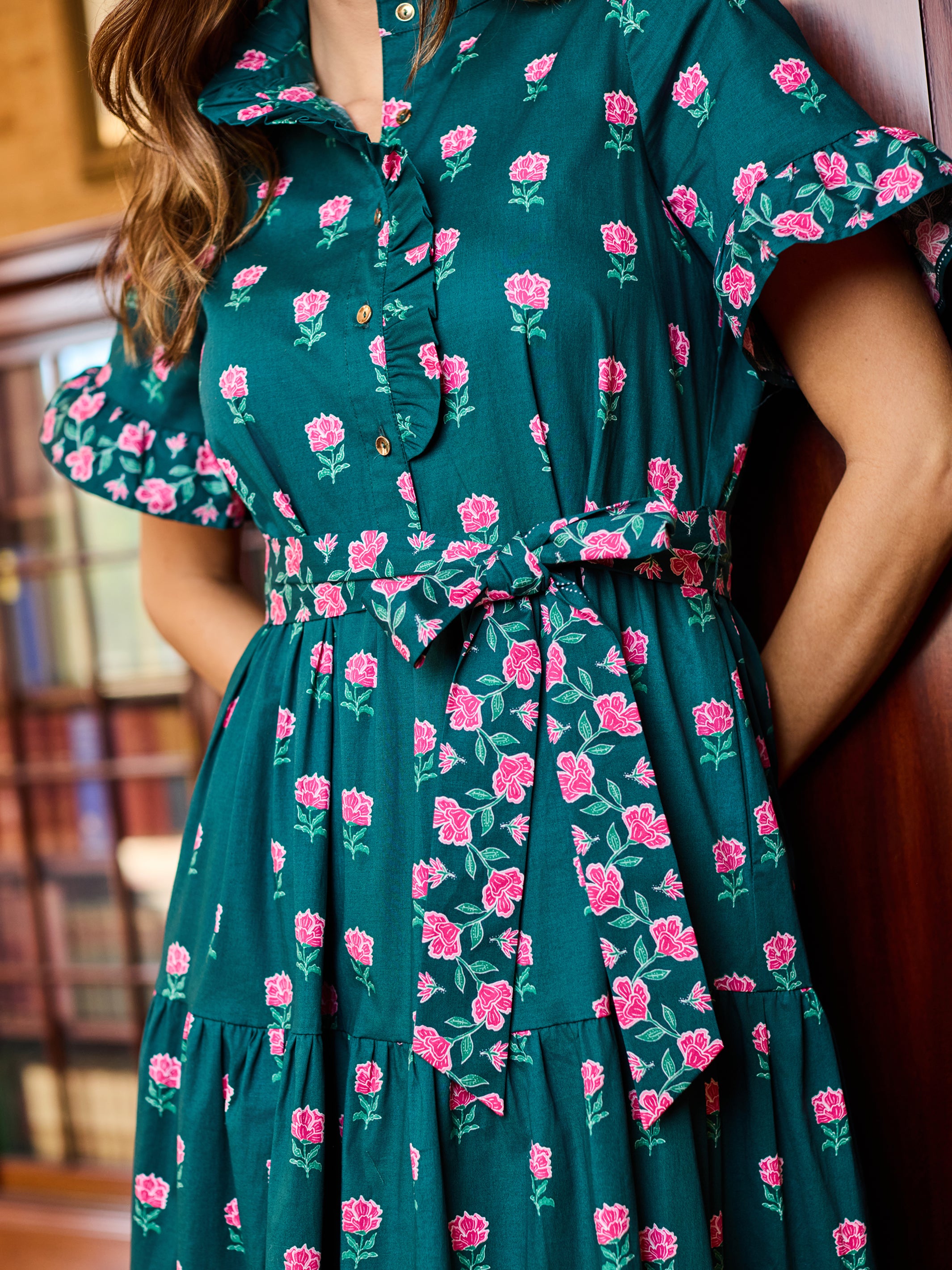 A woman wearing a dark teal green dress with a pink rose and green leaf floral print, featuring a ruffled collar, short flutter sleeves, and a matching waist tie.