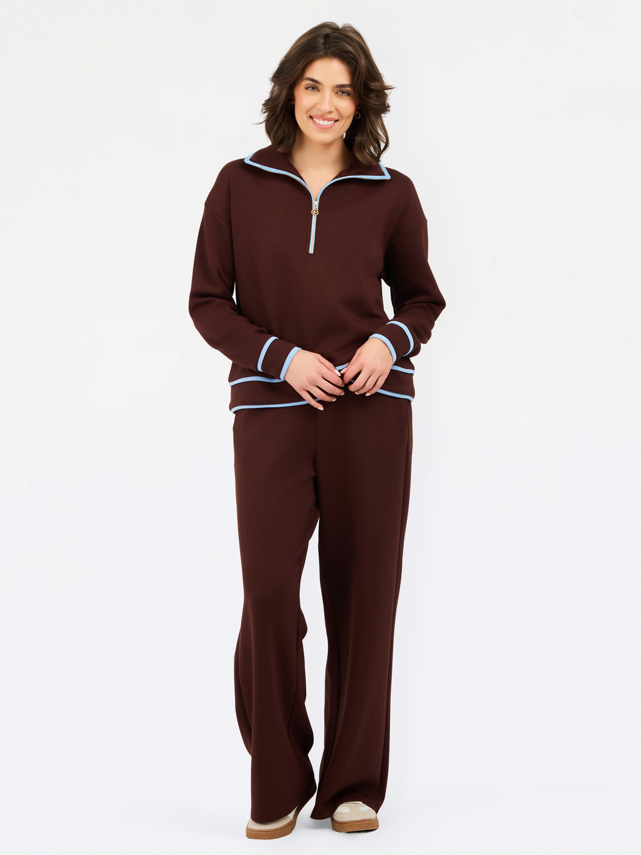 A woman modeling a matching two-piece lounge set in deep burgundy, featuring a quarter-zip long-sleeve top with light blue and white striped trim, and matching wide-leg pants.
