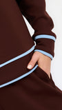 Close-up of a dark brown long-sleeve garment with light blue contrast piping along the sleeve cuff and hem, worn by a model whose hand is in a pocket, against a plain white background.