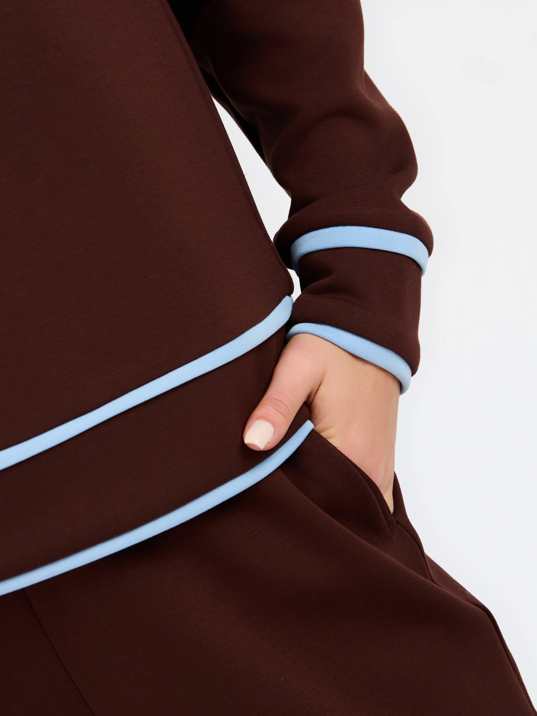 Close-up of a dark brown long-sleeve garment with light blue contrast piping along the sleeve cuff and hem, worn by a model whose hand is in a pocket, against a plain white background.