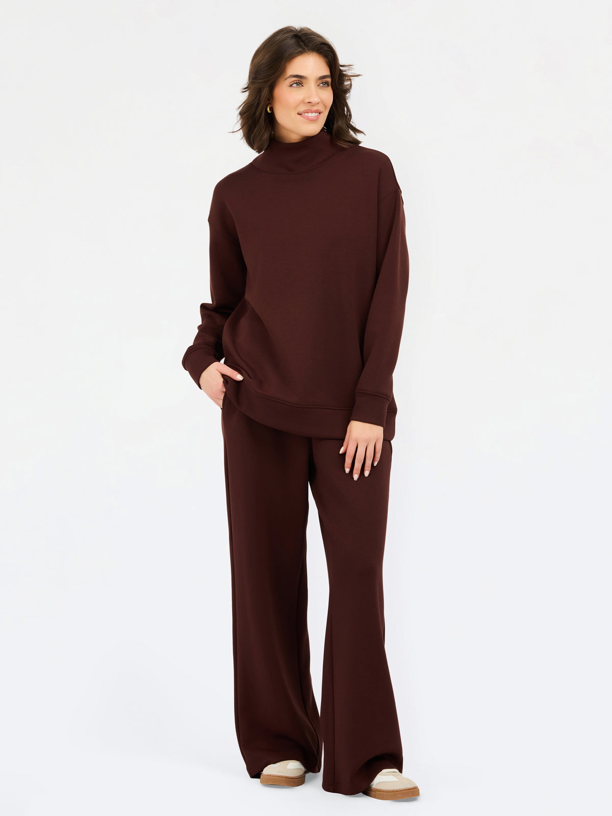 A woman wearing an oversized dark brown mock neck sweatshirt and matching sweatpants, photographed against a clean white studio background.