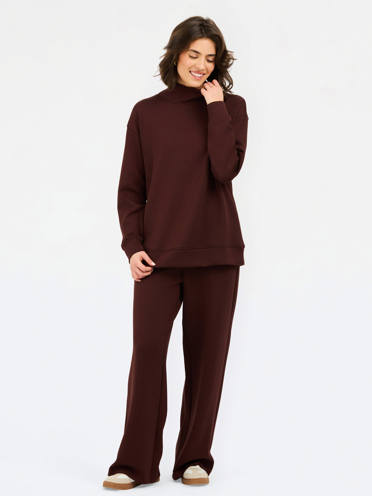 A woman wearing an oversized dark brown mock neck sweatshirt and matching sweatpants, photographed against a clean white studio background.