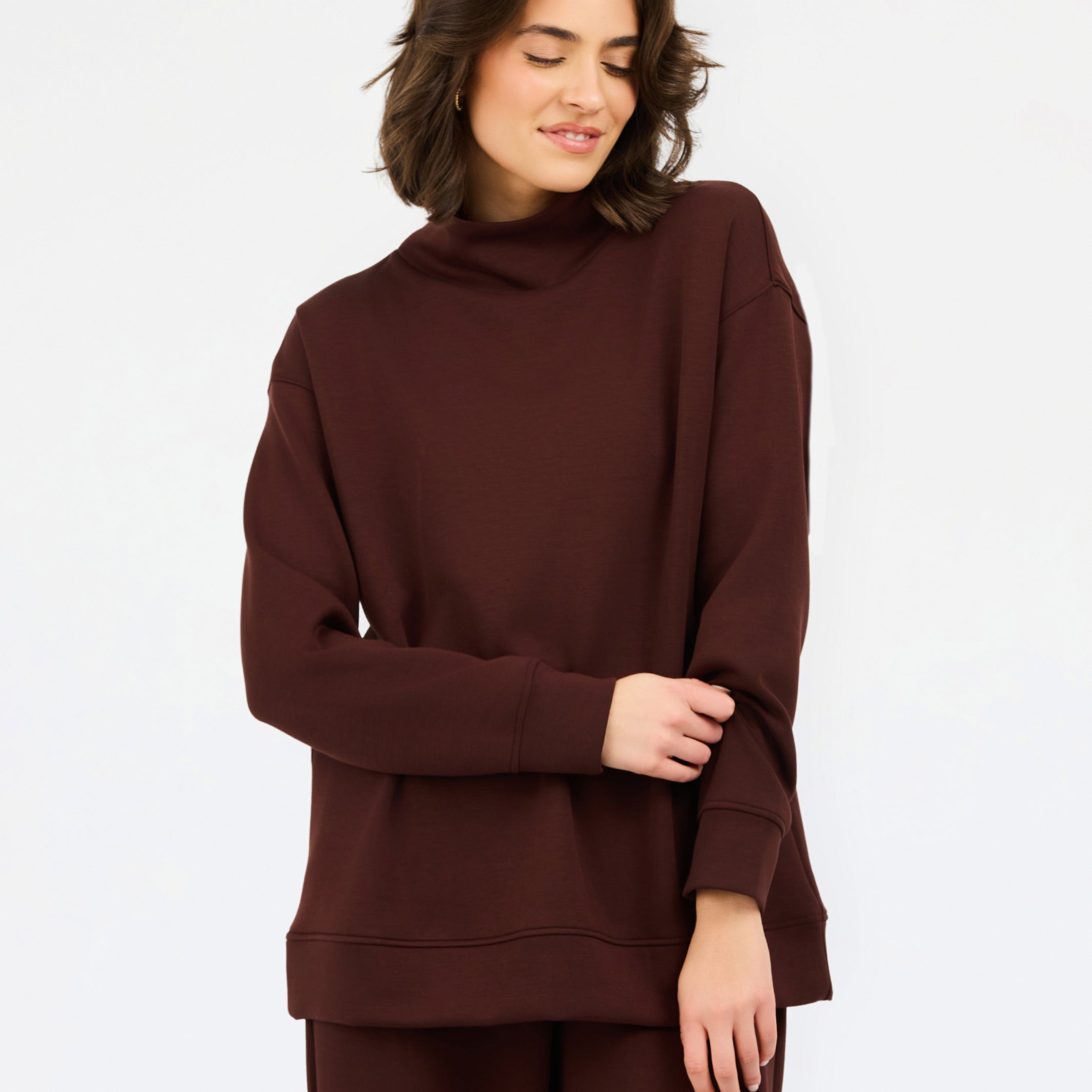 A woman wearing an oversized dark brown mock neck sweatshirt and matching sweatpants, photographed against a clean white studio background.