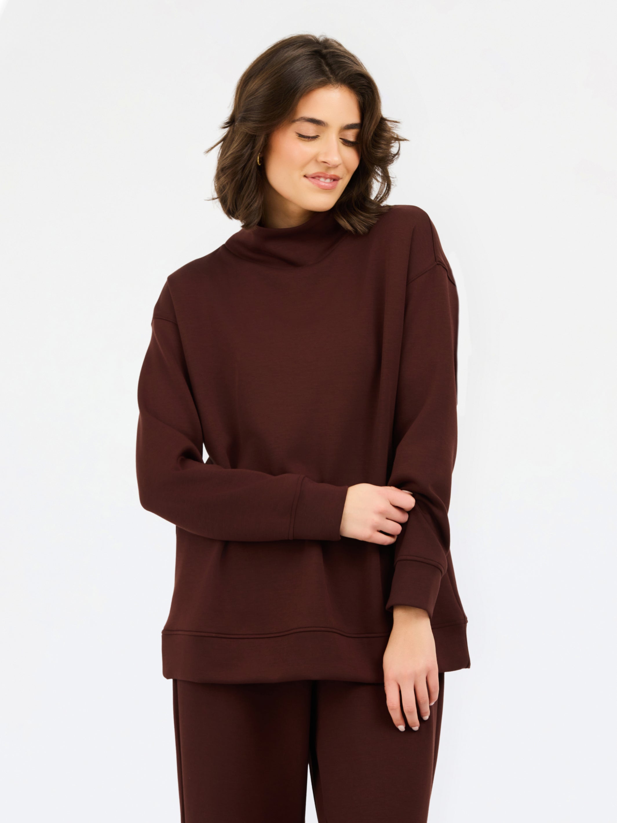 A woman wearing an oversized dark brown mock neck sweatshirt and matching sweatpants, photographed against a clean white studio background.