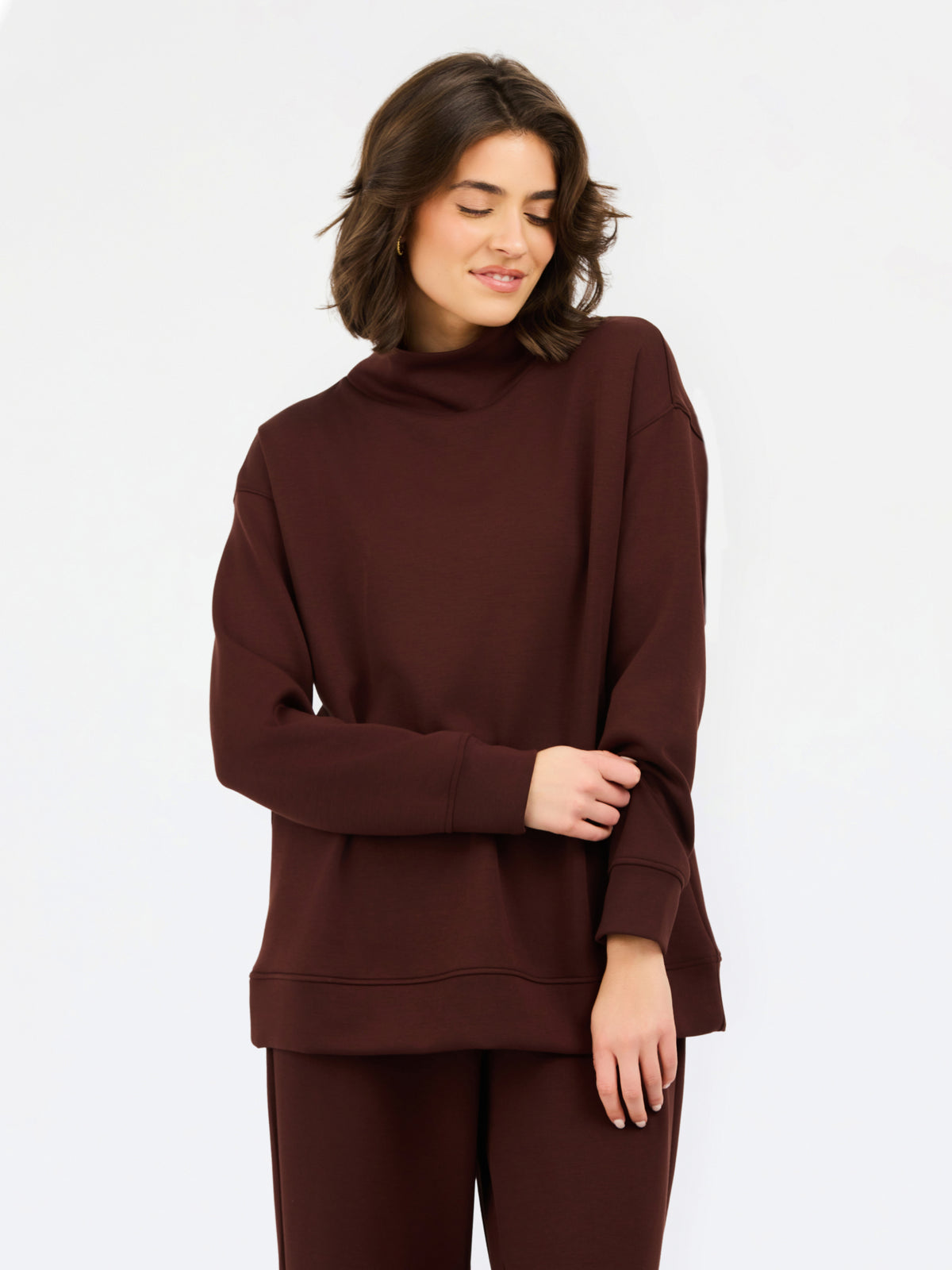 A woman wearing an oversized dark brown mock neck sweatshirt and matching sweatpants, photographed against a clean white studio background.
