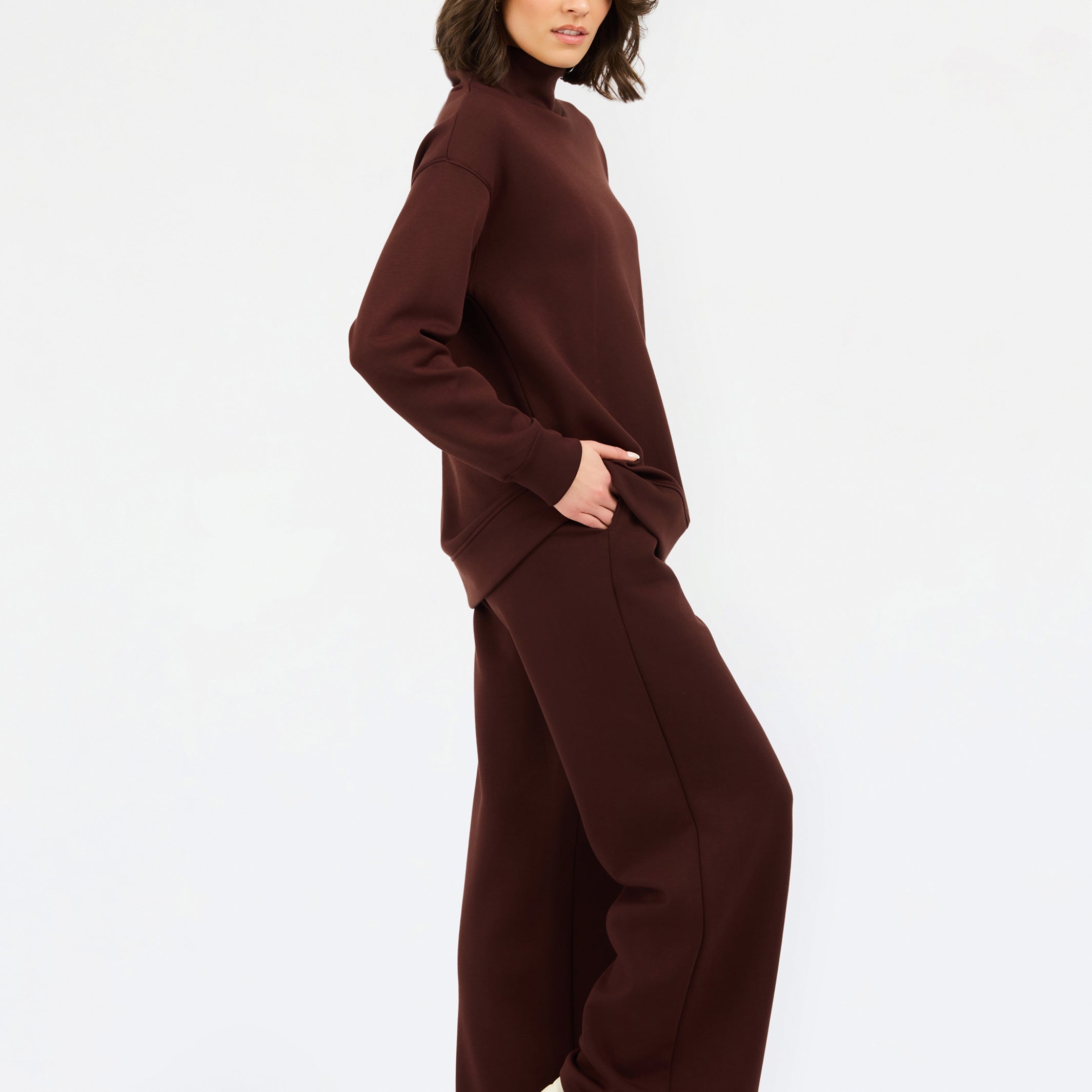 A woman wearing an oversized dark brown mock neck sweatshirt and matching sweatpants, photographed against a clean white studio background.