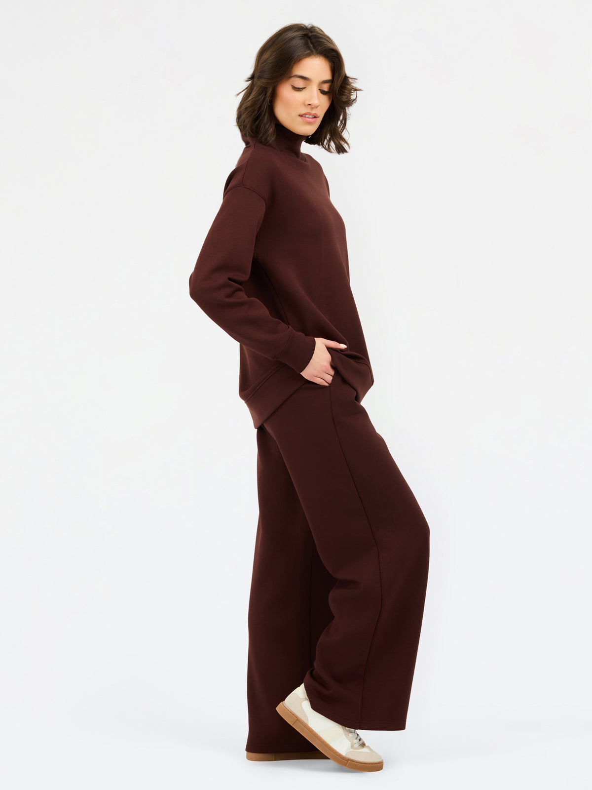 A woman wearing an oversized dark brown mock neck sweatshirt and matching sweatpants, photographed against a clean white studio background.