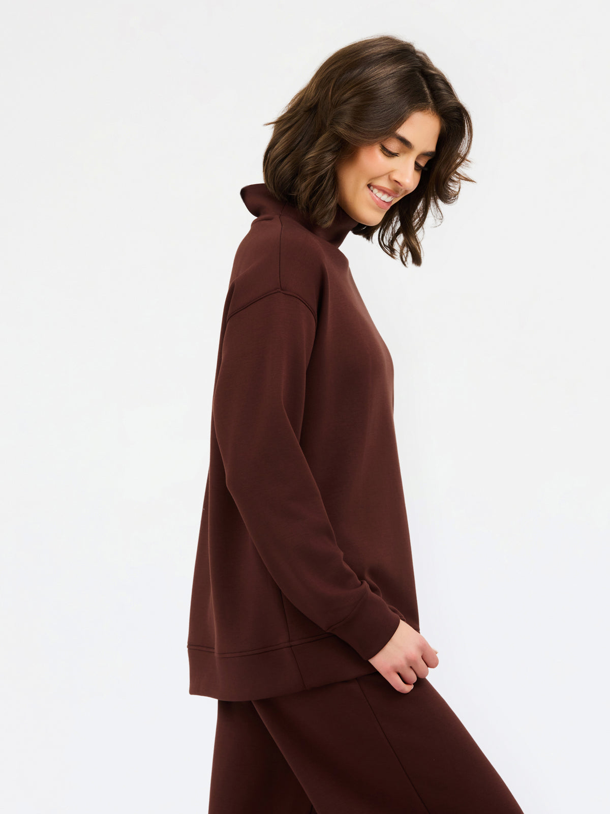 A woman wearing an oversized dark brown mock neck sweatshirt and matching sweatpants, photographed against a clean white studio background.