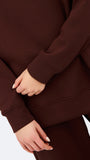 A woman wearing an oversized dark brown mock neck sweatshirt and matching sweatpants, photographed against a clean white studio background.