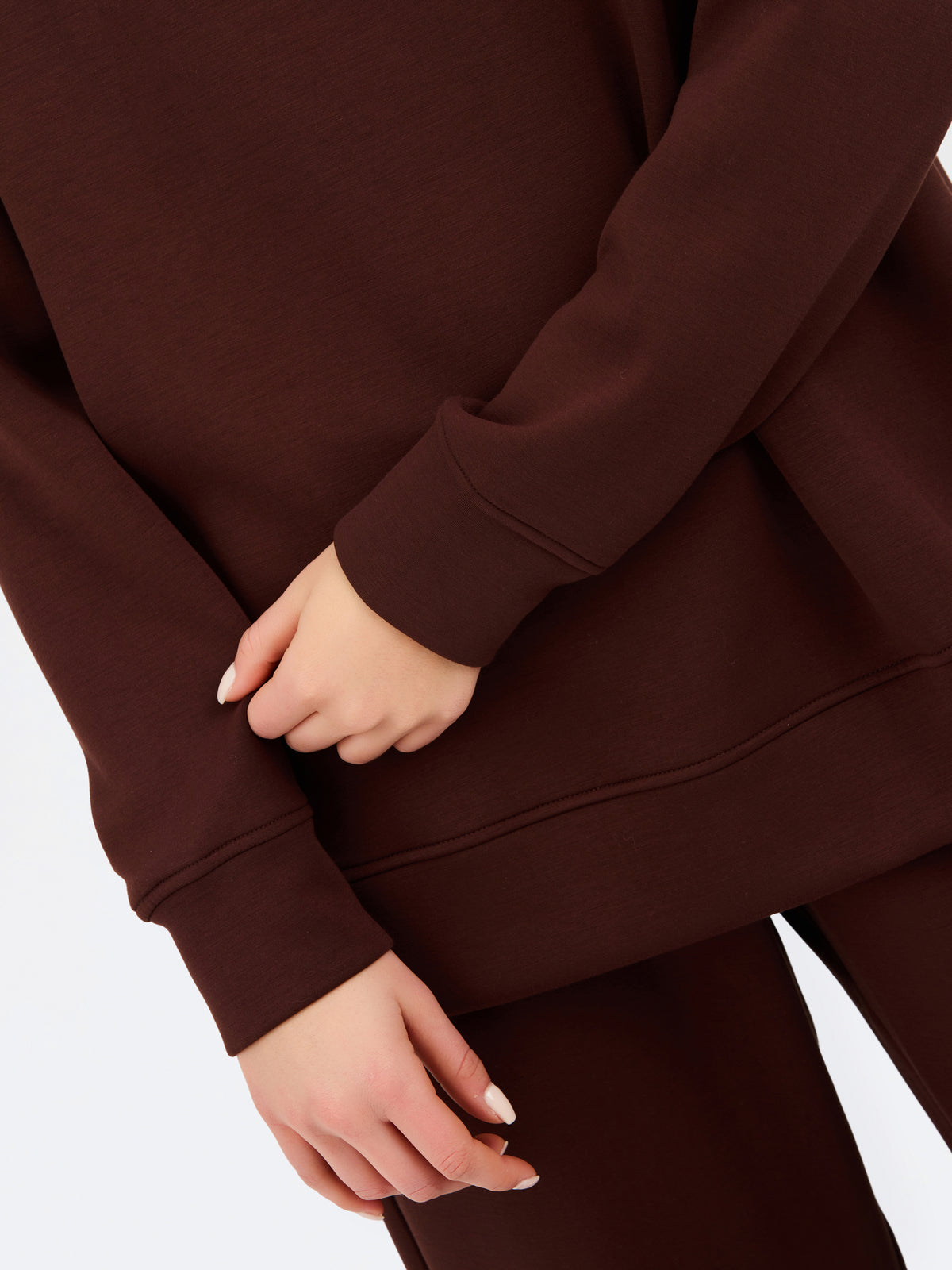 A woman wearing an oversized dark brown mock neck sweatshirt and matching sweatpants, photographed against a clean white studio background.
