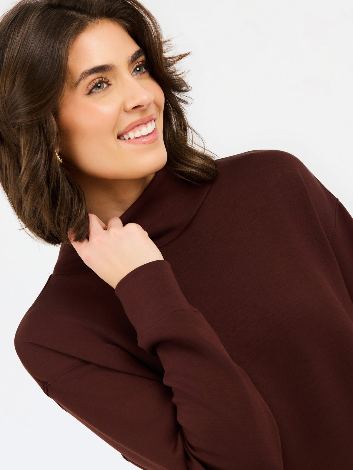 A woman wearing an oversized dark brown mock neck sweatshirt and matching sweatpants, photographed against a clean white studio background.