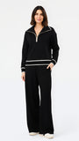 A woman modeling a black knit two-piece tracksuit with white contrast trim, featuring a half-zip pullover top and matching wide-leg pants, against a plain white studio background.
