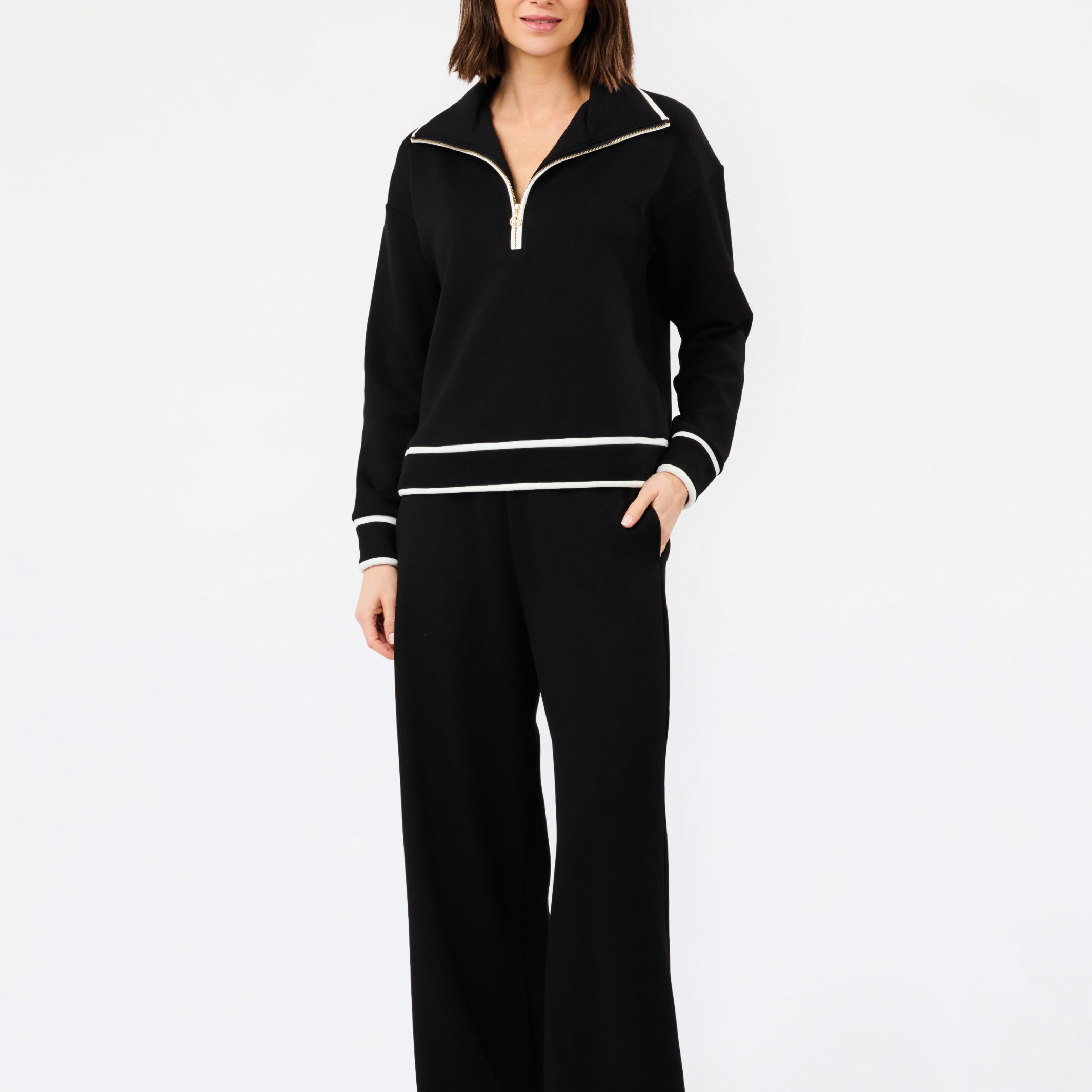 A woman modeling a black knit two-piece tracksuit with white contrast trim, featuring a half-zip pullover top and matching wide-leg pants, against a plain white studio background.