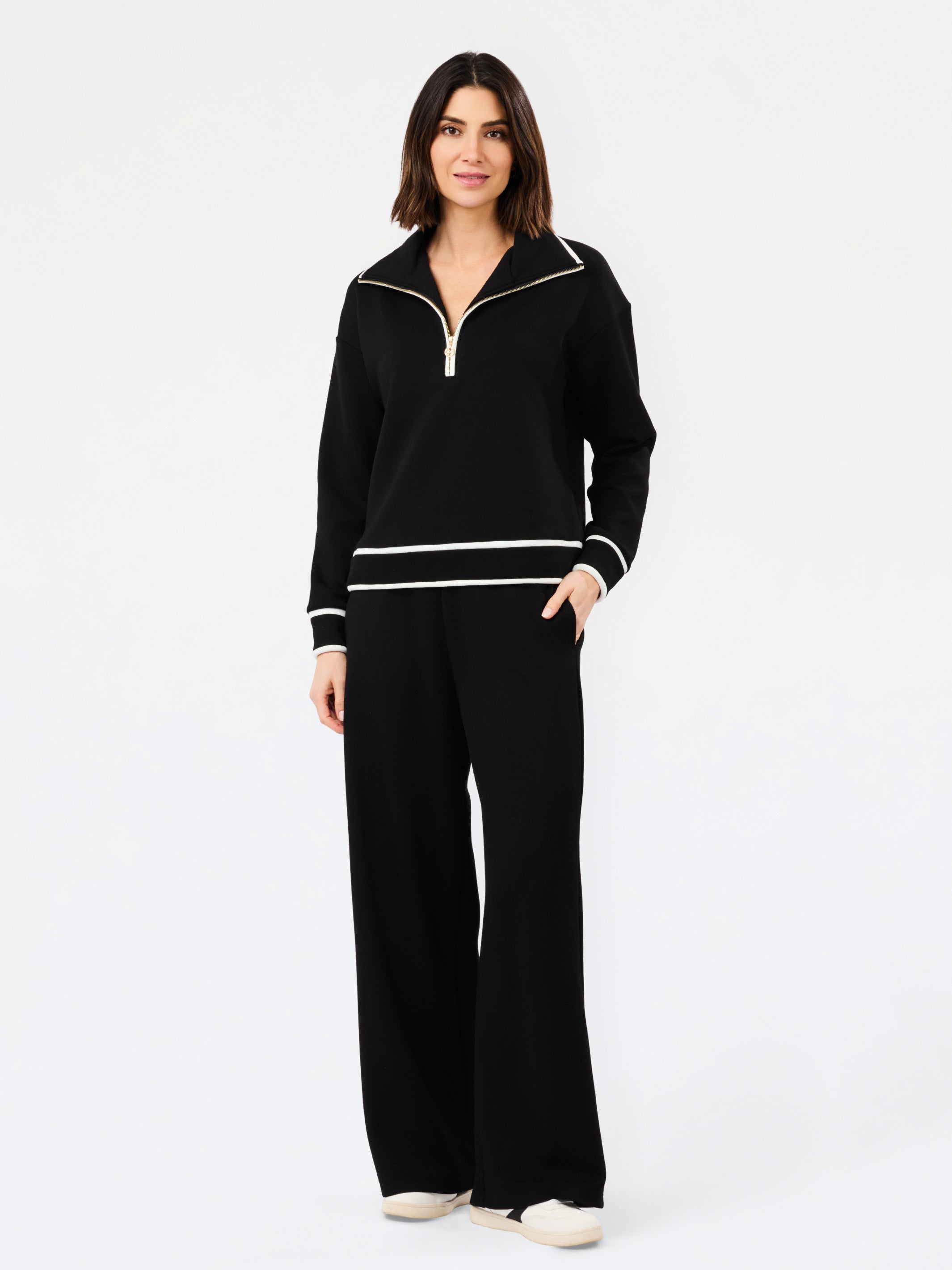 A woman modeling a black knit two-piece tracksuit with white contrast trim, featuring a half-zip pullover top and matching wide-leg pants, against a plain white studio background.
