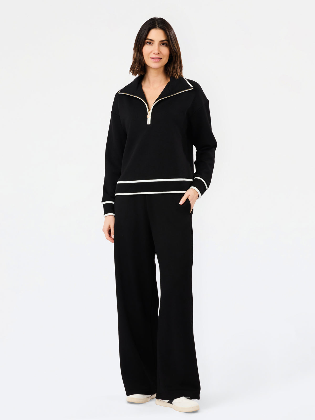 A woman modeling a black knit two-piece tracksuit with white contrast trim, featuring a half-zip pullover top and matching wide-leg pants, against a plain white studio background.
