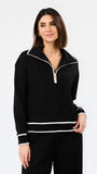 A woman modeling a black long-sleeve half-zip pullover with white contrast trim and a gold zipper pull, paired with black pants, against a plain white studio background.