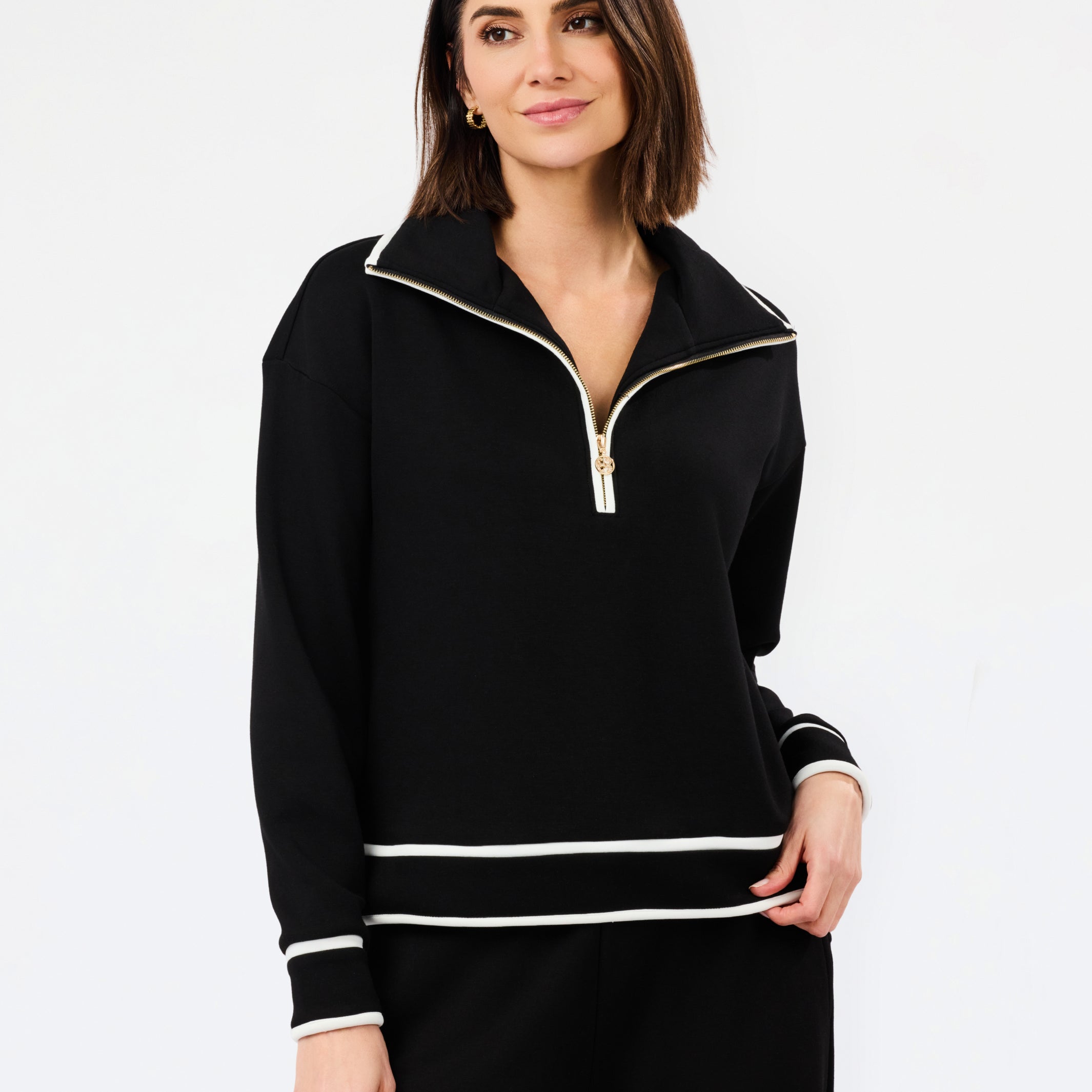 A woman modeling a black long-sleeve half-zip pullover with white contrast trim and a gold zipper pull, paired with black pants, against a plain white studio background.