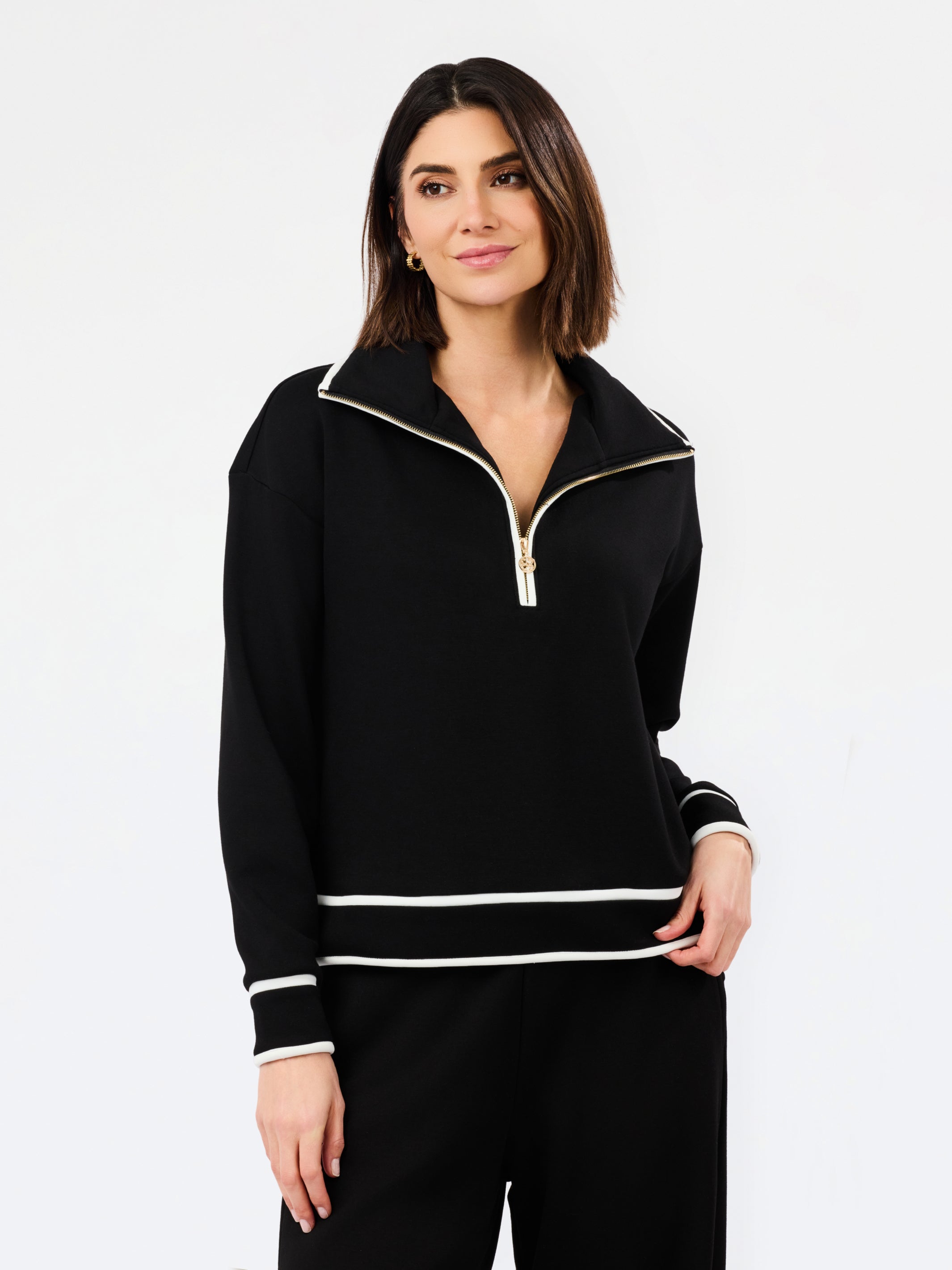 A woman modeling a black long-sleeve half-zip pullover with white contrast trim and a gold zipper pull, paired with black pants, against a plain white studio background.