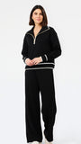 A woman modeling a black knit two-piece tracksuit with white contrast trim, featuring a half-zip pullover top and matching wide-leg pants, against a plain white studio background.