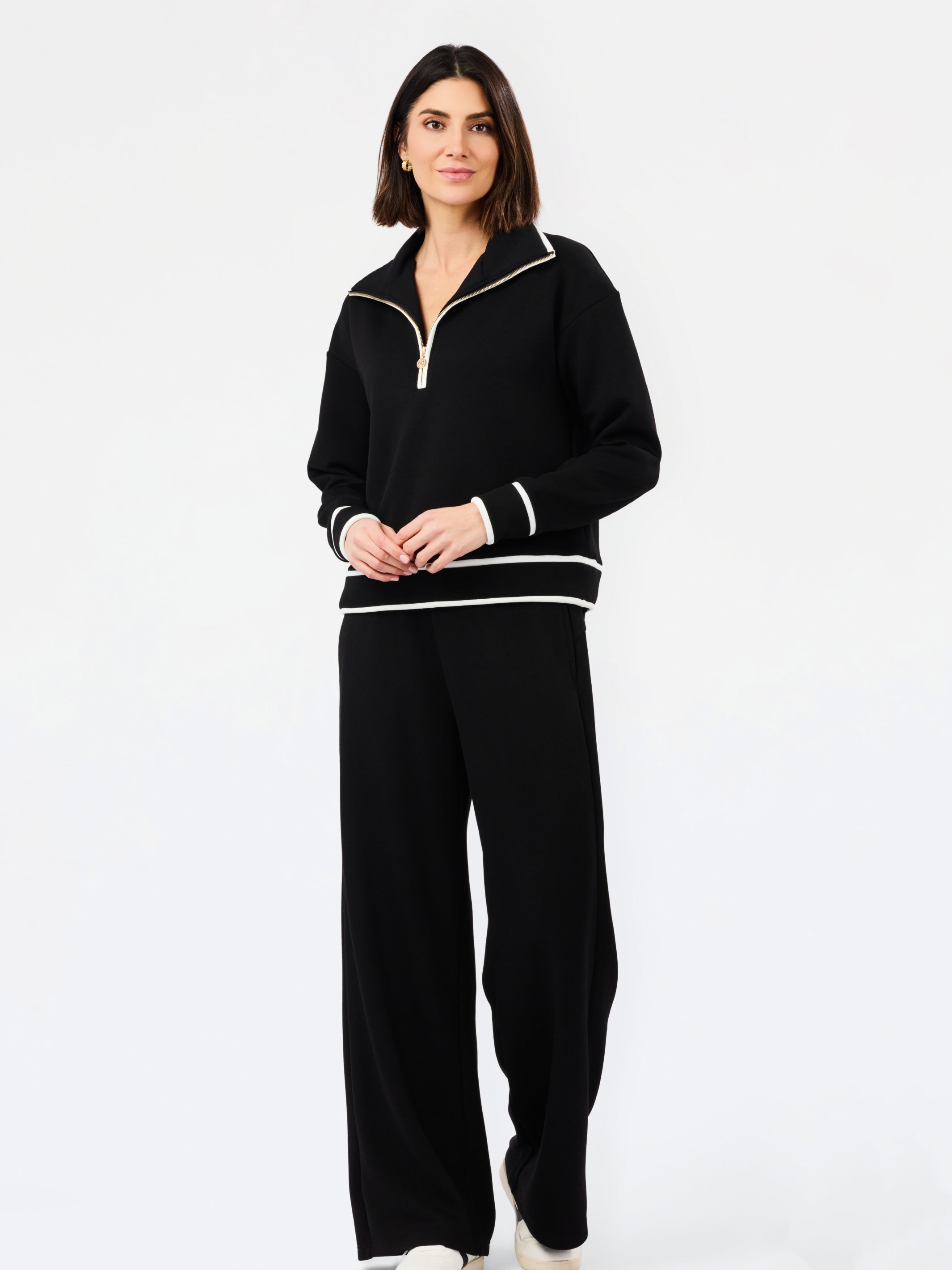 A woman modeling a black knit two-piece tracksuit with white contrast trim, featuring a half-zip pullover top and matching wide-leg pants, against a plain white studio background.
