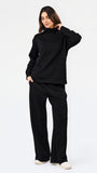 A woman modeling a black two-piece loungewear set consisting of a long-sleeve mock neck sweatshirt and matching wide-leg sweatpants, paired with white slip-on shoes, against a plain white studio background.