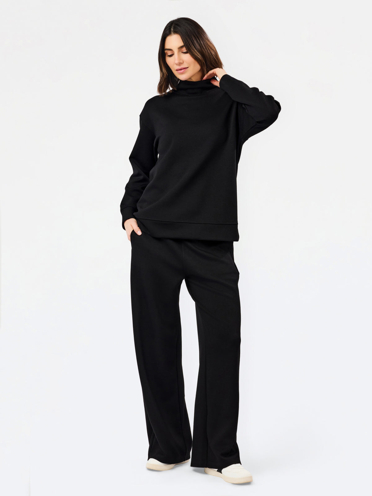 A woman modeling a black two-piece loungewear set consisting of a long-sleeve mock neck sweatshirt and matching wide-leg sweatpants, paired with white slip-on shoes, against a plain white studio background.
