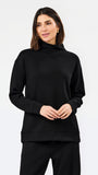 A woman modeling a black long-sleeve turtleneck sweatshirt and matching black pants against a plain white studio background.