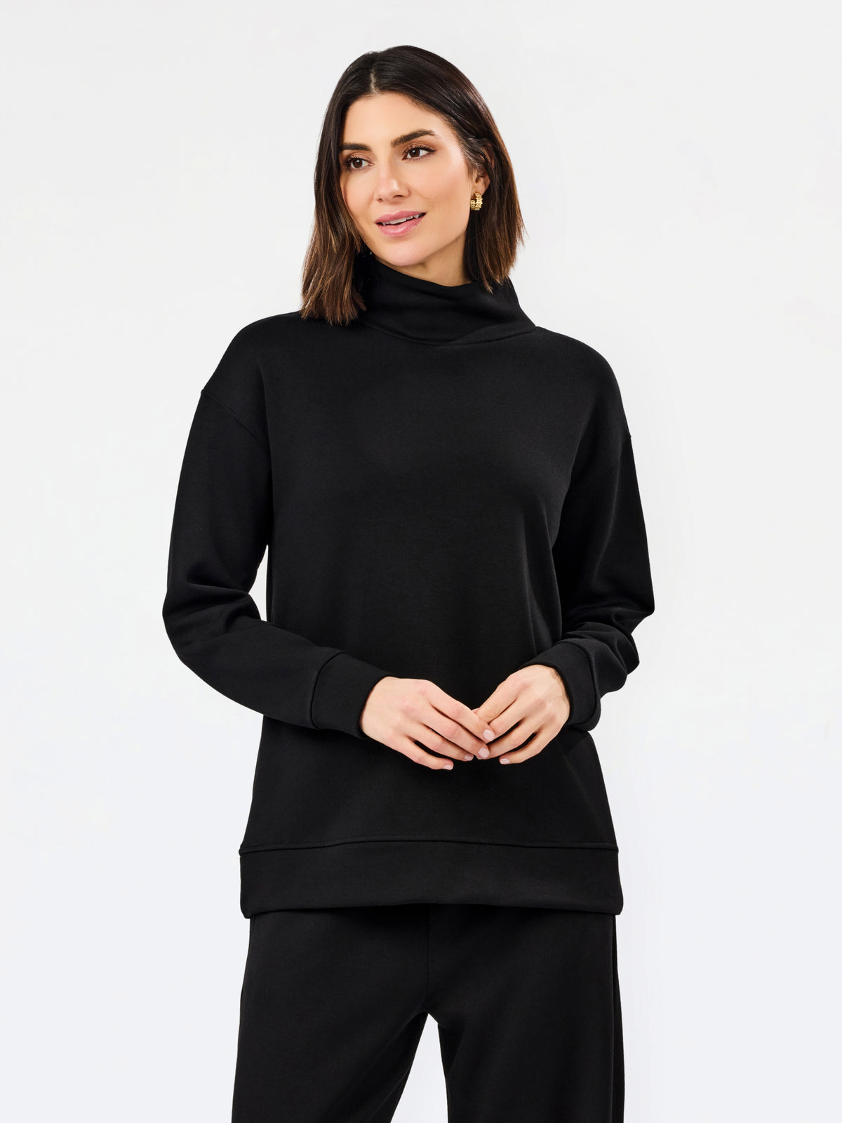 A woman modeling a black long-sleeve turtleneck sweatshirt and matching black pants against a plain white studio background.
