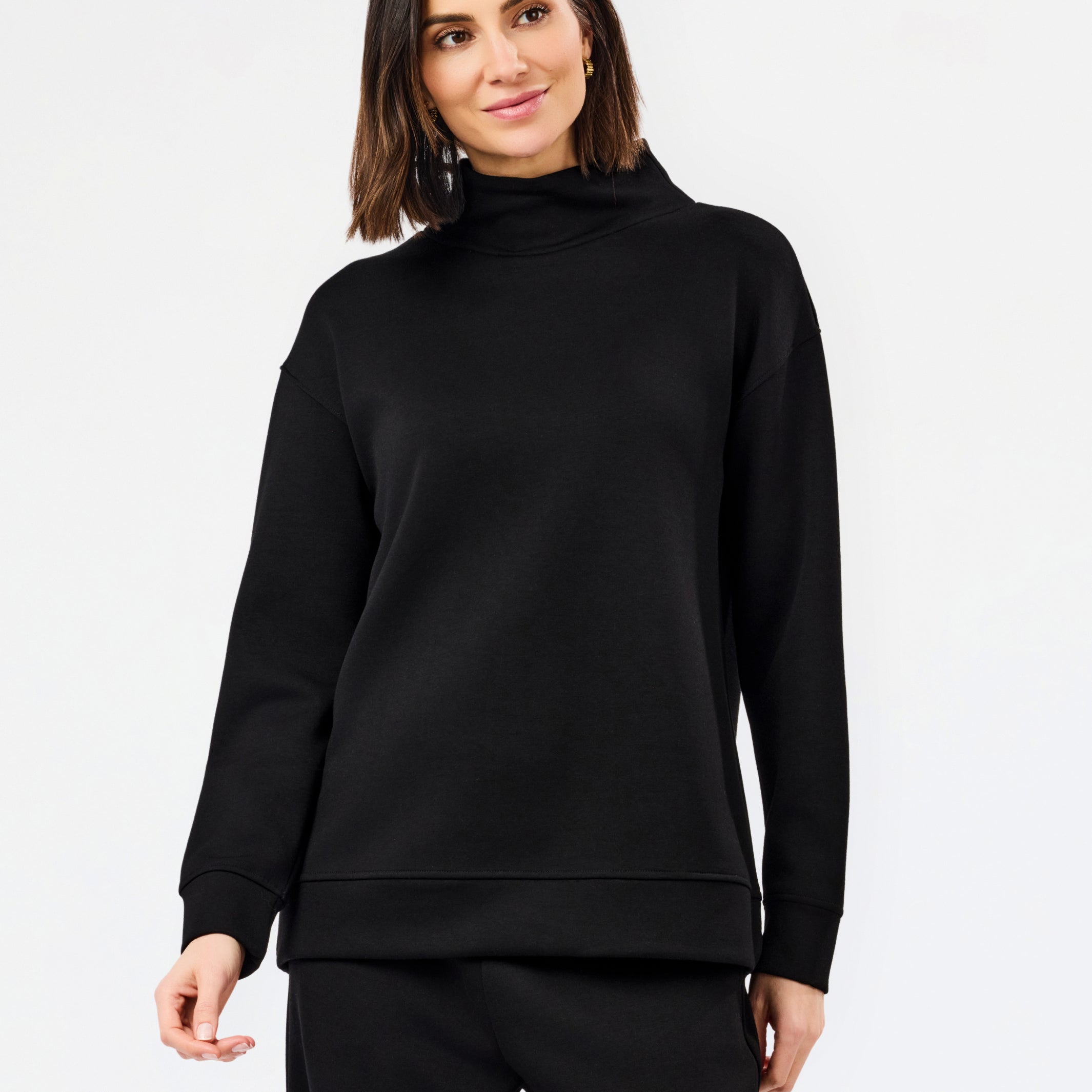 A woman modeling a black long-sleeve mock neck sweatshirt and matching black pants against a plain white studio background.