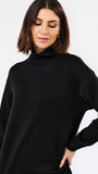 A woman wearing an oversized black long-sleeve turtleneck top, photographed against a clean light gray studio background.