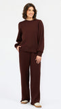 A woman modeling a matching dark brown knit lounge set consisting of a crewneck sweatshirt and wide-leg pants, paired with beige slip-on shoes, against a plain white studio background.